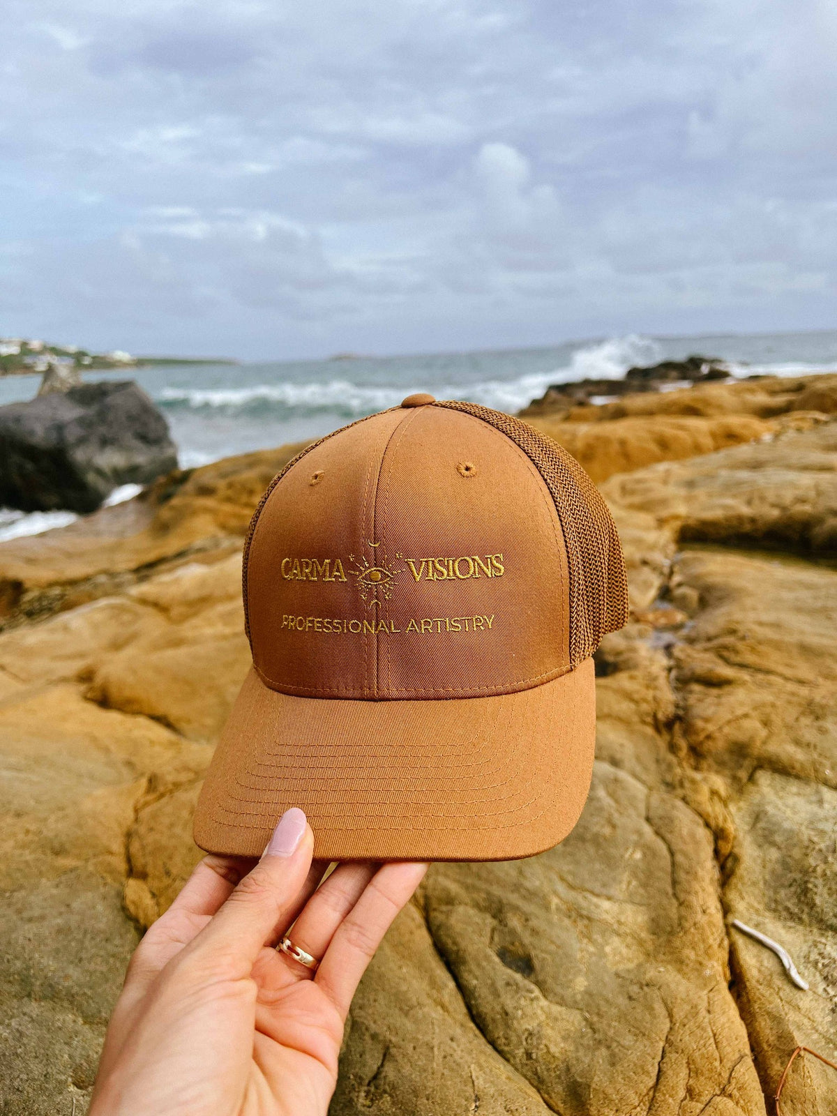 Carma Visions trucker cap in caramel, held on a rocky beach